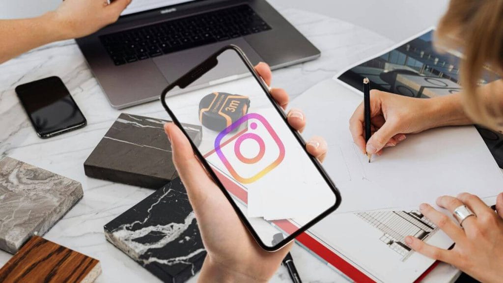 Mão a segurar telemóvel com o logótipo do Instagram, sobre uma mesa de trabalho de arquitetura com plantas, esquisso e amostras de materiais.
