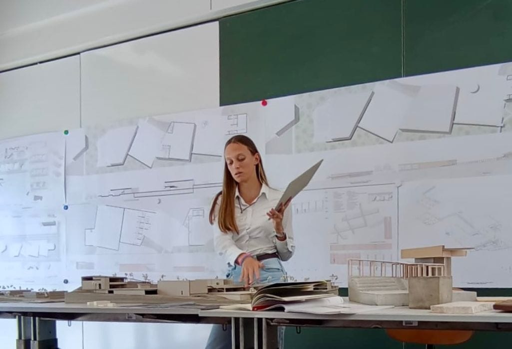 Debora Madile, fundadora da A Arquiteta da tua Marca, a apresentar um projeto de arquitetura na faculdade, com maquetes sobre a mesa e desenhos de arquitetura afixados na parede.